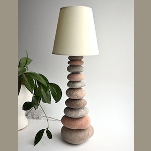 May include: A distinctive table lamp featuring a base made of stacked stones and a cream lampshade. The base is constructed from smooth stones of different sizes and hues, offering a natural, textured look. The lamp is a decorative lighting element.