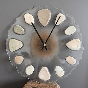 May include: A unique wall clock with a clear, wavy-edged face. The clock features a central brown design and is adorned with smooth, light-colored stones. Black clock hands indicate the time. The clock is mounted on a wooden base.