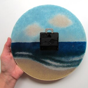 Pu&ograve; includere: Un orologio rotondo in vetro con una scena di spiaggia. Il quadrante dell'orologio presenta un cielo blu con nuvole bianche, un mare blu con onde bianche e una spiaggia sabbiosa. Il meccanismo dell'orologio &egrave; visibile sul retro.