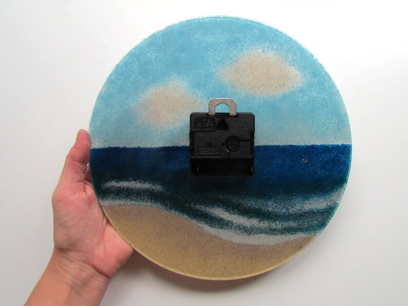 Pu&ograve; includere: Un orologio rotondo in vetro con una scena di spiaggia. Il quadrante dell'orologio presenta un cielo blu con nuvole bianche, un mare blu con onde bianche e una spiaggia sabbiosa. Il meccanismo dell'orologio &egrave; visibile sul retro.