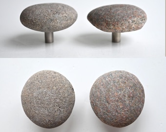 May include: Two cabinet knobs featuring natural, rounded river stones as the front face. Each stone has a textured, speckled grey and reddish-brown surface. The stones are securely attached to a cylindrical, metallic silver-toned metal base.