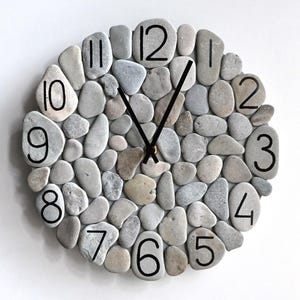 May include: A circular wall clock constructed from smooth, grey and beige stones. Black numerals mark the hours, and black hands indicate the time. The clock is mounted on a white wall.