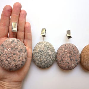 May include: Four round, smooth stones of varying shades of pink, gray, and tan, each featuring a silver-colored metal wire clip attached to the top. The stones have a natural, speckled texture and are arranged in a horizontal row.