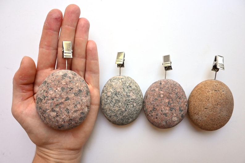 May include: Four round, smooth stones of varying shades of pink, gray, and tan, each featuring a silver-colored metal wire clip attached to the top. The stones have a natural, speckled texture and are arranged in a horizontal row.