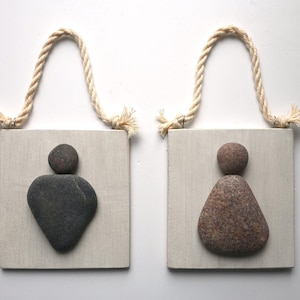 May include: Two wooden restroom signs with rope hangers. Each sign features a stone figure. One is dark gray, the other is brown. The signs are mounted on light gray wooden plaques.