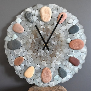 May include: A round clock with a unique design. The clock face is made of small, light gray stones and larger, colorful pebbles. Black clock hands point to the time. The clock is a decorative item.