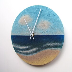 Pu&ograve; includere: Un orologio rotondo con un design a paesaggio marino blu e bianco. Il quadrante dell'orologio &egrave; fatto di sabbia e presenta lancette bianche e un centro dorato.