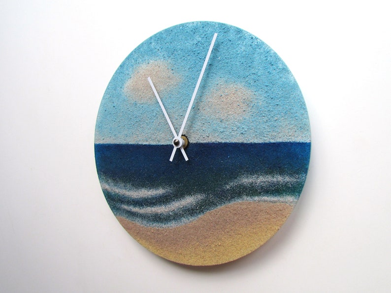 Pu&ograve; includere: Un orologio rotondo con un design a paesaggio marino blu e bianco. Il quadrante dell'orologio &egrave; fatto di sabbia e presenta lancette bianche e un centro dorato.