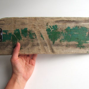 Op de afbeelding: Een rechthoekige houten plank met een verweerde afwerking, met vlekken groene verf en twee metalen haken. Het hout vertoont zichtbare nerven en textuur, met een verweerde uitstraling. De haken zijn zilverkleurig en aan het hout bevestigd.