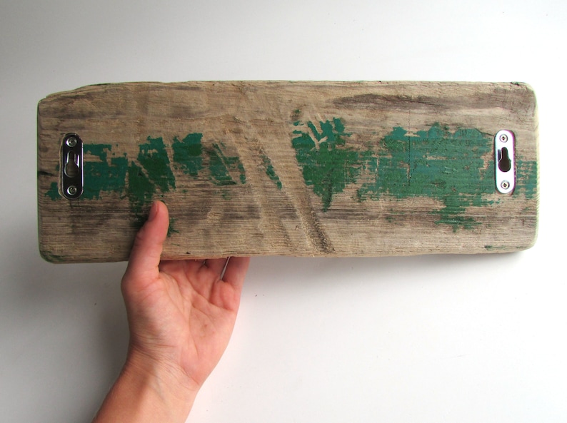 Op de afbeelding: Een rechthoekige houten plank met een verweerde afwerking, met vlekken groene verf en twee metalen haken. Het hout vertoont zichtbare nerven en textuur, met een verweerde uitstraling. De haken zijn zilverkleurig en aan het hout bevestigd.