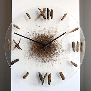 May include: A clear, round wall clock with black hands and Roman numerals made of small, dark brown wood pieces. The clock face has a textured, brown center and is mounted on a white wall.