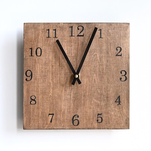May include: Square wooden wall clock with black numerals and hands. The clock face is a light brown colour, and the hands point to approximately 11:06. The clock is mounted on a white wall.