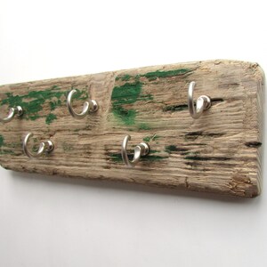 Op de afbeelding: Een rustieke sleutelhouder gemaakt van een verweerde houten plank met zes zilverkleurige haken. Het hout heeft een natuurlijke, verouderde uitstraling met groene verfplekken. Een uniek en functioneel item voor in huis.