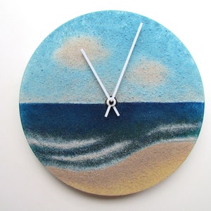 Pu&ograve; includere: Un orologio rotondo con una scena di spiaggia dipinta su di esso. Il quadrante dell'orologio &egrave; diviso in un cielo blu con nuvole bianche, un mare blu con onde bianche e una spiaggia color sabbia. Le lancette dell'orologio sono bianche.