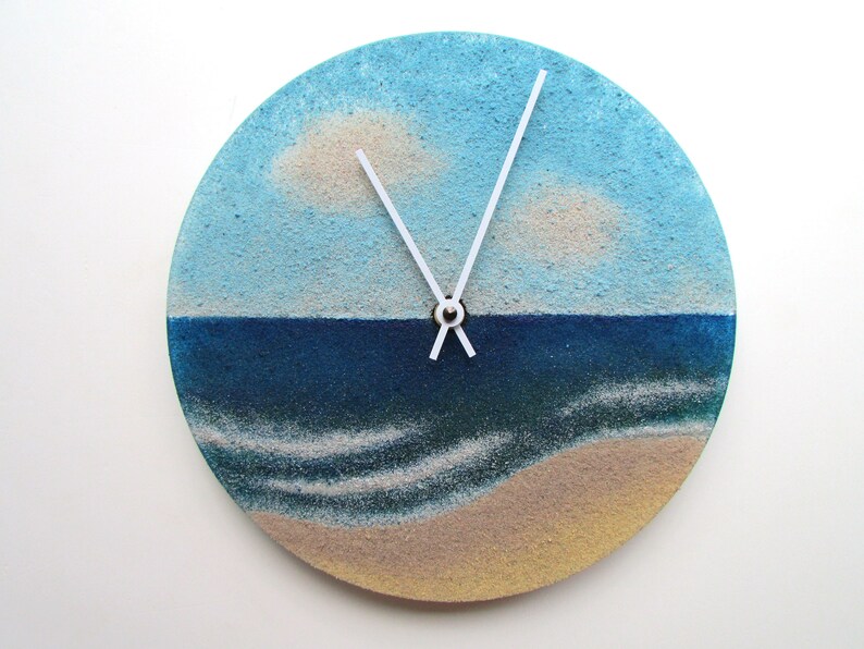 Pu&ograve; includere: Un orologio rotondo con una scena di spiaggia dipinta su di esso. Il quadrante dell'orologio &egrave; diviso in un cielo blu con nuvole bianche, un mare blu con onde bianche e una spiaggia color sabbia. Le lancette dell'orologio sono bianche.