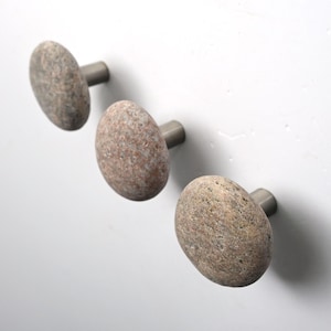 Può includere: Tre pomelli da parete con superfici in pietra liscia e arrotondata, maculate nei toni del marrone chiaro e del grigio, fissate a basi metalliche cilindriche. I pomelli sono disposti in diagonale, evidenziando la texture naturale e i toni terra.