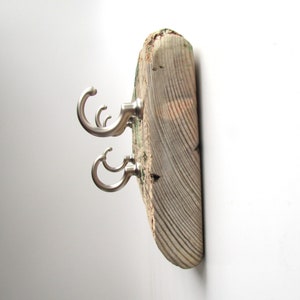 Op de afbeelding: Een rustieke wandkapstok gemaakt van een verweerde houten plank. Het heeft drie geborsteld nikkel haken, elk met een gebogen ontwerp. Het hout heeft zichtbare nerven en natuurlijke kleurvariaties, wat bijdraagt aan de charme.