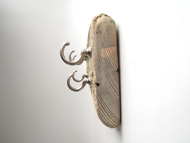 Op de afbeelding: Een rustieke wandkapstok gemaakt van een verweerde houten plank. Het heeft drie geborsteld nikkel haken, elk met een gebogen ontwerp. Het hout heeft zichtbare nerven en natuurlijke kleurvariaties, wat bijdraagt aan de charme.