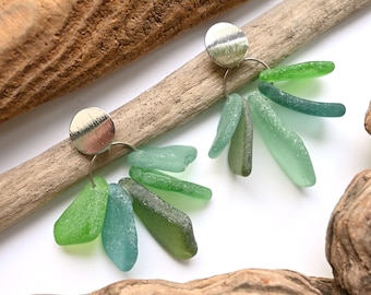 May include: A pair of handcrafted earrings featuring silver-toned circular studs and dangling sea glass pieces in shades of green and blue. The earrings are displayed on a piece of driftwood, showcasing their natural and organic aesthetic.