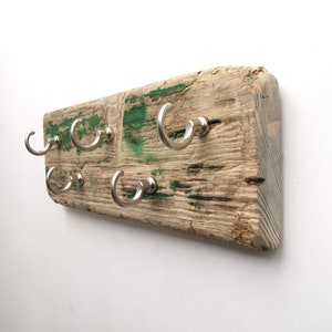 Op de afbeelding: Een rustieke houten wandkapstok met zes zilverkleurige metalen haken. Het hout is verweerd met zichtbare nerven en groene verfplekken. Een decoratief en functioneel item voor de hal of slaapkamer.