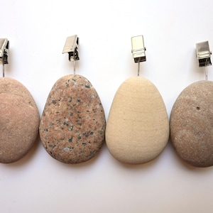 May include: Four smooth, oval-shaped stones arranged in a row, each topped with a silver-coloured metal alligator clip attached by a thin wire. The stones vary in colour and texture, including shades of tan, speckled pink, beige, and light brown.