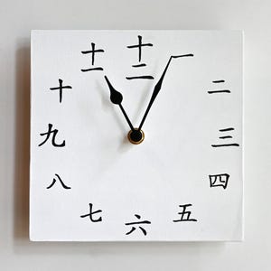 以下が含まれることがあります： 白い正方形の壁掛け時計。文字盤には1から12までの日本語の数字が円状に配置されています。時計には黒い針が2本あり、11時と1時の位置を指しています。中央には小さな金色のキャップが付いています。