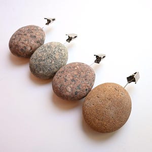 May include: Four smooth, oval-shaped river stones in varying shades of brown, gray, and tan, each featuring a silver-toned metal alligator clip attached to a thin wire stem extending from the top of the stone for holding cards or photos.