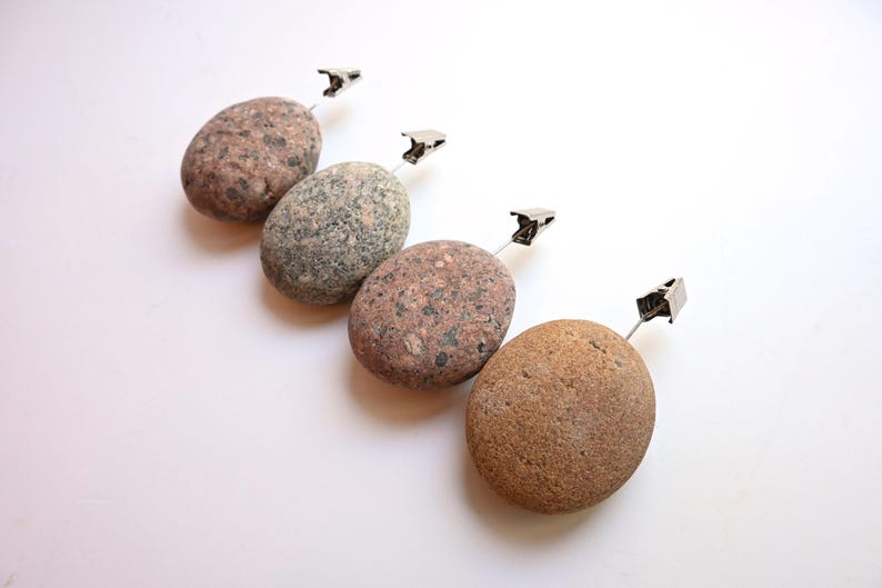 May include: Four smooth, oval-shaped river stones in varying shades of brown, gray, and tan, each featuring a silver-toned metal alligator clip attached to a thin wire stem extending from the top of the stone for holding cards or photos.