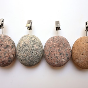 May include: Four round, smooth stones used as photo or card holders, each topped with a silver-colored metal alligator clip. The stones feature natural textures and varied earth tones, including speckled pink, grey, and light brown variations.
