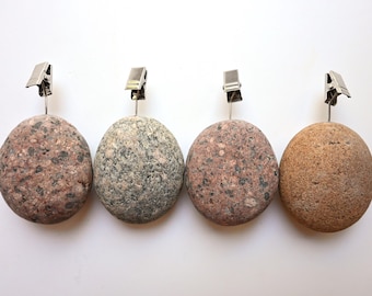 May include: Four round, smooth stones used as photo or card holders, each topped with a silver-colored metal alligator clip. The stones feature natural textures and varied earth tones, including speckled pink, grey, and light brown variations.