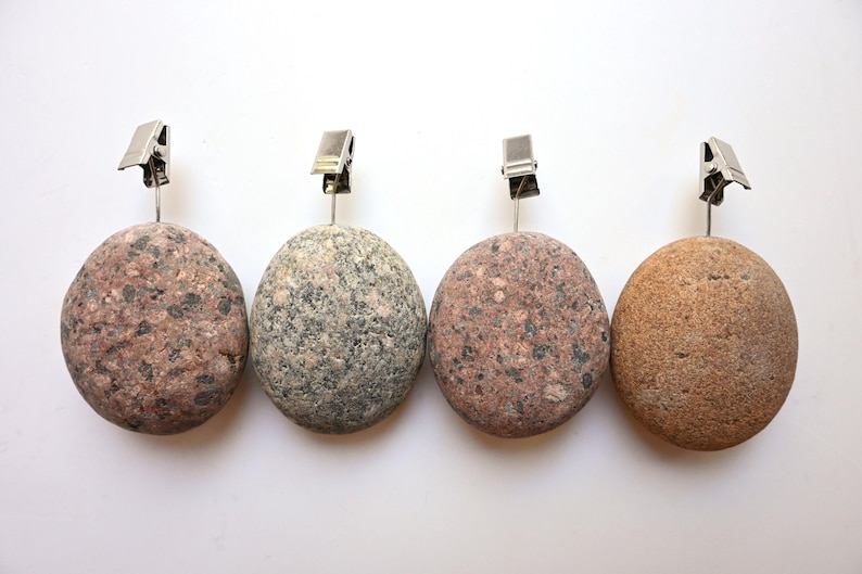 May include: Four round, smooth stones used as photo or card holders, each topped with a silver-colored metal alligator clip. The stones feature natural textures and varied earth tones, including speckled pink, grey, and light brown variations.