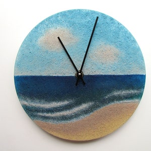 Pu&ograve; includere: Un orologio rotondo con una scena di spiaggia dipinta su di esso. Il quadrante dell'orologio &egrave; blu e bianco, con una spiaggia sabbiosa in basso. Le lancette dell'orologio sono nere.