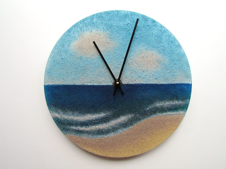 Pu&ograve; includere: Un orologio rotondo con una scena di spiaggia dipinta su di esso. Il quadrante dell'orologio &egrave; blu e bianco, con una spiaggia sabbiosa in basso. Le lancette dell'orologio sono nere.