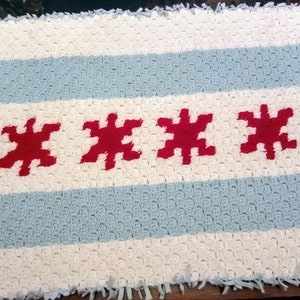 May include: A crocheted blanket featuring the Chicago flag. The blanket is white with blue and red stripes and three red stars.
