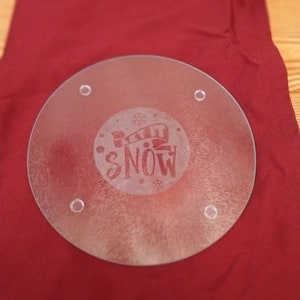 May include: A clear glass round cutting board with a frosted design that says "Let it Snow" in a circle with snowflakes. The cutting board has four small holes around the edge.