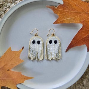 May include: A pair of ghost-shaped dangle earrings crafted from small white and cream-colored beads with two black beads forming the eyes. Each earring features a gold-toned metal hook for attachment, creating a hanging design with a fringe edge.