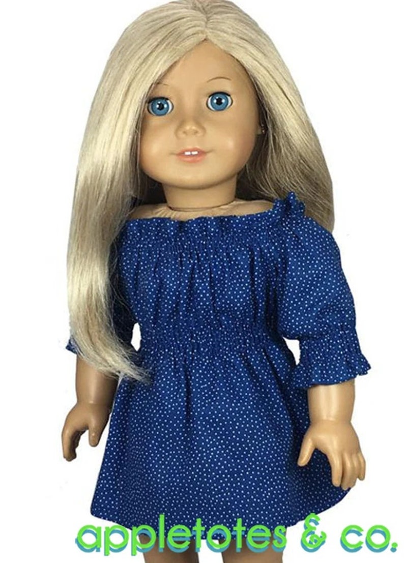 May include: A doll wearing a blue and white polka dot off-the-shoulder dress with a ruffled neckline and 3/4 sleeves. The doll has blonde hair and blue eyes. The dress is made for 18-inch dolls. The image includes the text "appletotes & co."