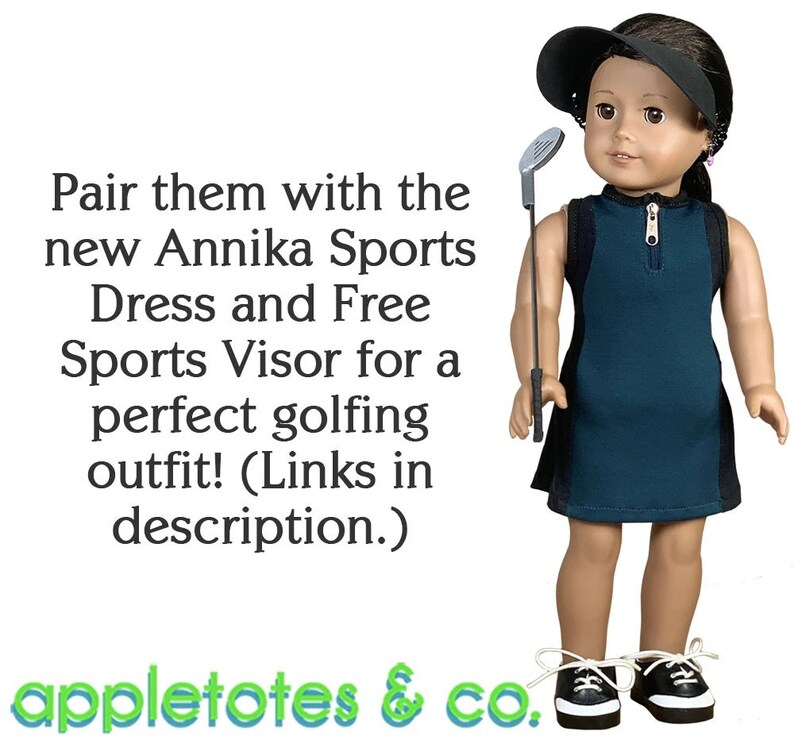 May include: A doll wearing a teal sleeveless dress with a black stripe down the side, a black visor, and black and white sneakers. The doll is holding a golf club. The text "Pair them with the new Annika Sports Dress and Free Sports Visor for a perfect golfing outfit! (Links in description.) appletotes & co." is at the bottom of the image.