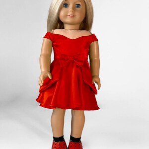 May include: A doll with blonde hair and blue eyes wearing a red dress with a bow and matching red shoes with black accents. The doll is standing against a white background.