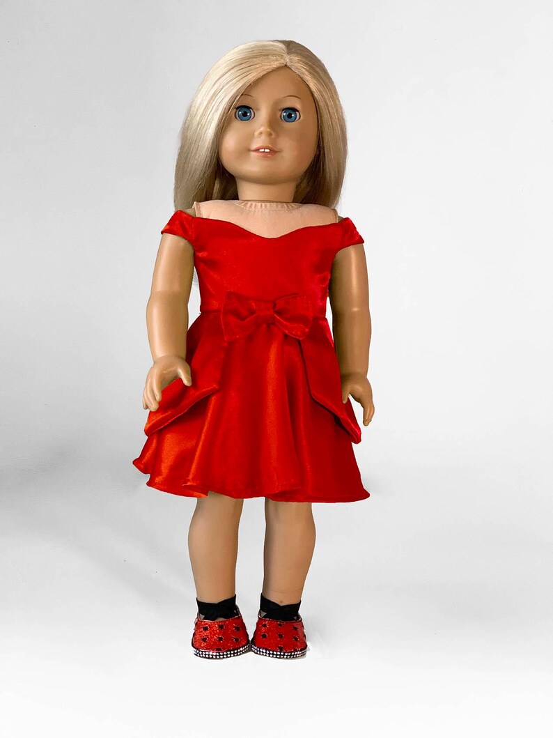 May include: A doll with blonde hair and blue eyes wearing a red dress with a bow and matching red shoes with black accents. The doll is standing against a white background.