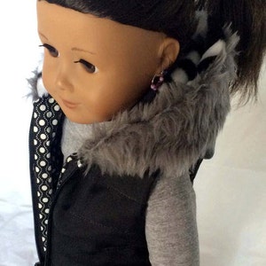 May include: A black puffer vest with a gray faux fur hood and a white and black polka dot lining. The vest has a zipper closure and is worn by a doll with brown hair.