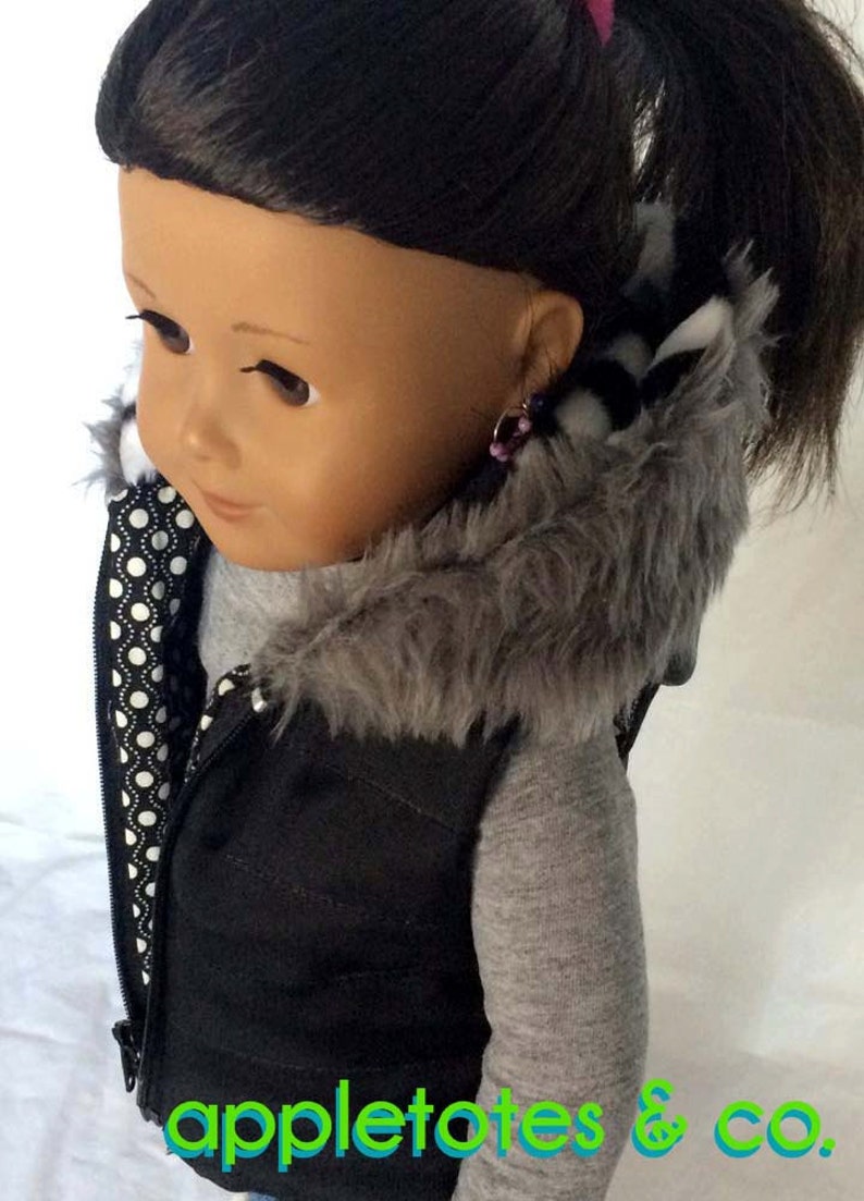 May include: A black puffer vest with a gray faux fur hood and a white and black polka dot lining. The vest has a zipper closure and is worn by a doll with brown hair.