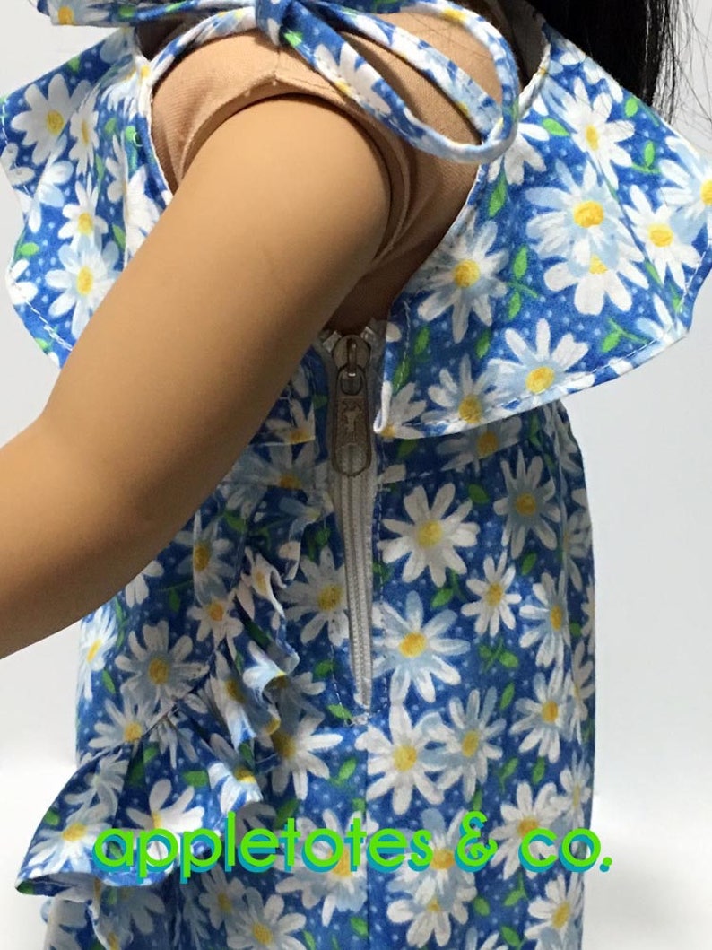 May include: A blue and white daisy print dress with a white ruffle trim and a zipper closure. The dress has a tie at the neck and is made for a doll.