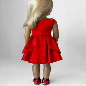 May include: A doll wearing a bright red dress with a pleated skirt and off-the-shoulder neckline. The doll has blonde hair and is wearing red and black sequined shoes. The doll is standing against a white background.