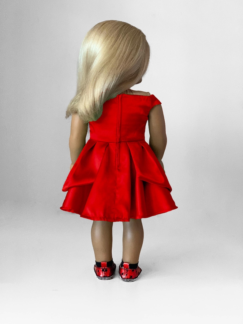 May include: A doll wearing a bright red dress with a pleated skirt and off-the-shoulder neckline. The doll has blonde hair and is wearing red and black sequined shoes. The doll is standing against a white background.