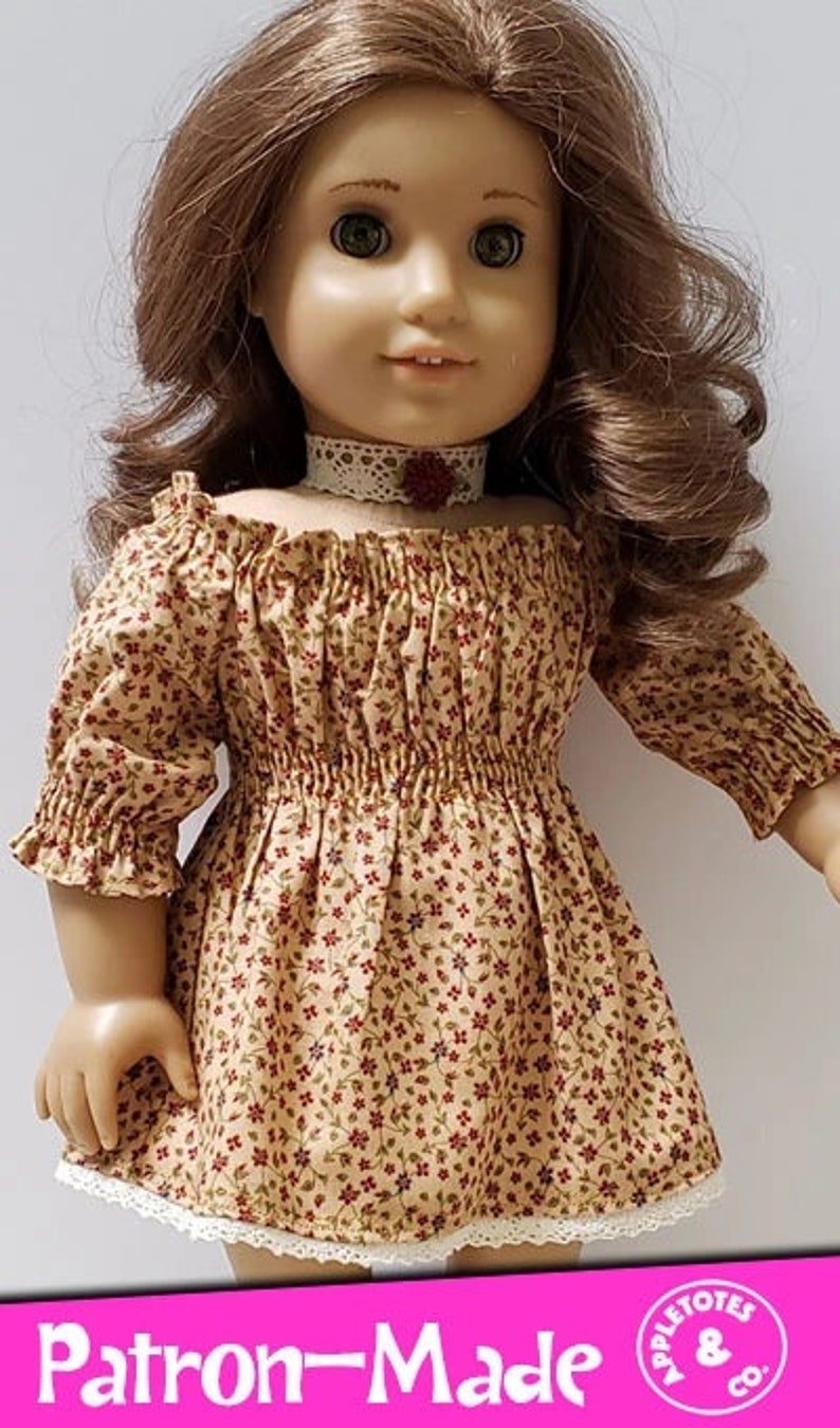 May include: A doll wearing a brown and white floral off-the-shoulder dress with a white lace trim. The dress has a gathered waist and 3/4 sleeves. The doll is wearing a white lace choker with a red flower accent. The doll has long brown hair.