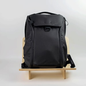 May include: A black backpack with a zippered front pocket and adjustable straps. The backpack is sitting on a wooden stand.