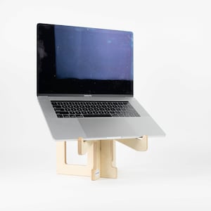 May include: A laptop computer on a wooden stand. The laptop is silver and has a black screen. The stand is made of light wood and has a simple design.