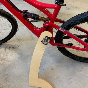 May include: A red mountain bike with a wooden bike stand. The bike has a black suspension fork and a black seat post. The stand is made of light wood and has a curved shape.