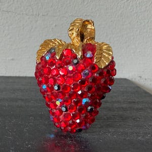 May include: A strawberry-shaped brooch, encrusted with vibrant red rhinestones. The brooch features a gold-toned stem and leaves, adding a touch of elegance. The sparkling design makes it a unique accessory.